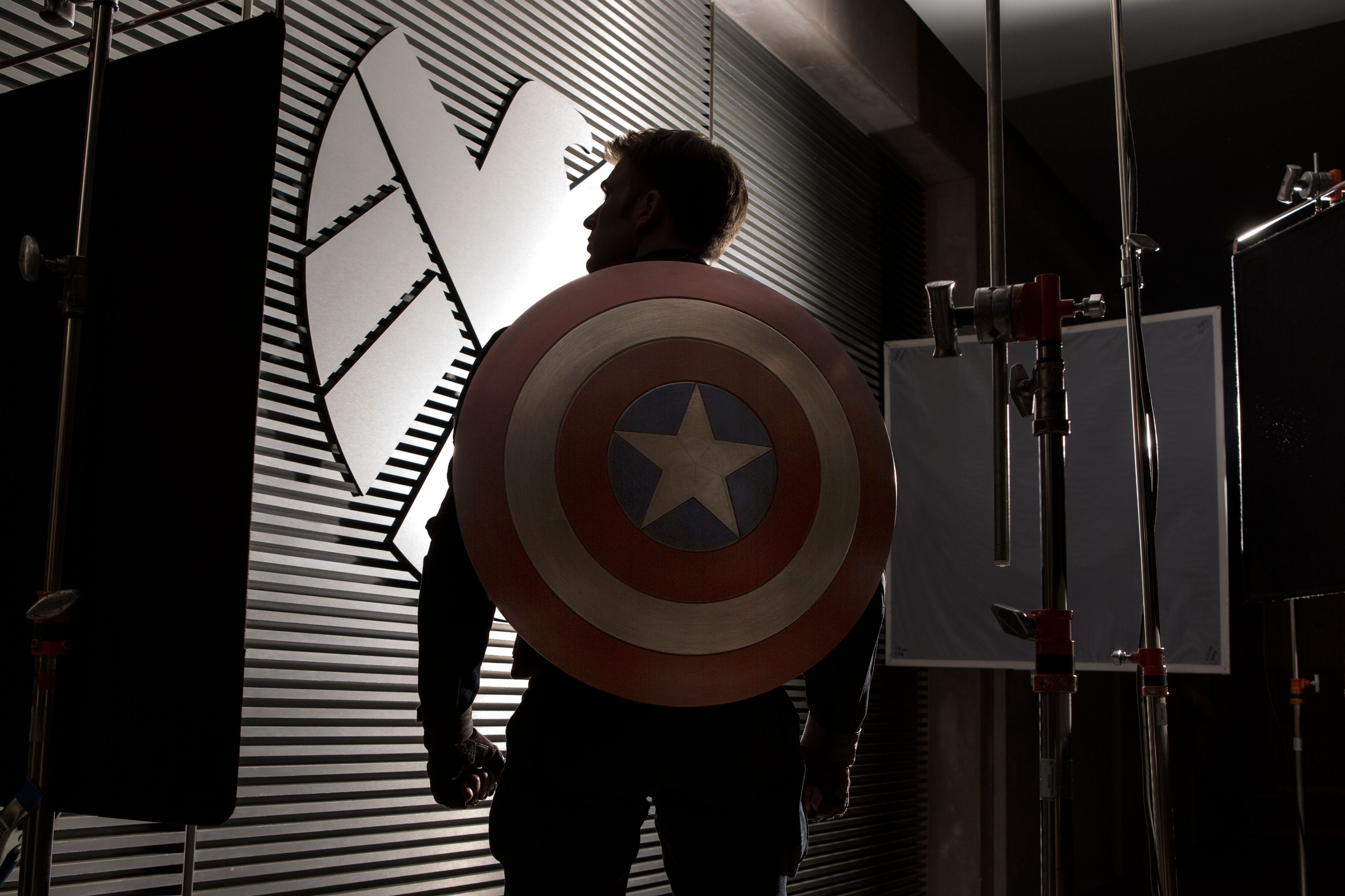 Capitão América de costas, à sua frente uma parede com o logo da Shield
