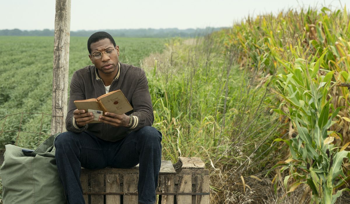 Lovecraft Country (2020). Alone in a green field is Tick Freeman, sitting on a crate reading a book besides a green sack. He is wearing round glasses and a brown jacket over a white t-shirt and dark blue jeans. He looks concentrated on what he is reading.